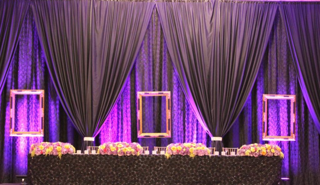 A table covered with a black tablecloth and floral arrangements sits in front of a backdrop of purple curtains with three empty gold frames hanging, all lit with purple lighting.