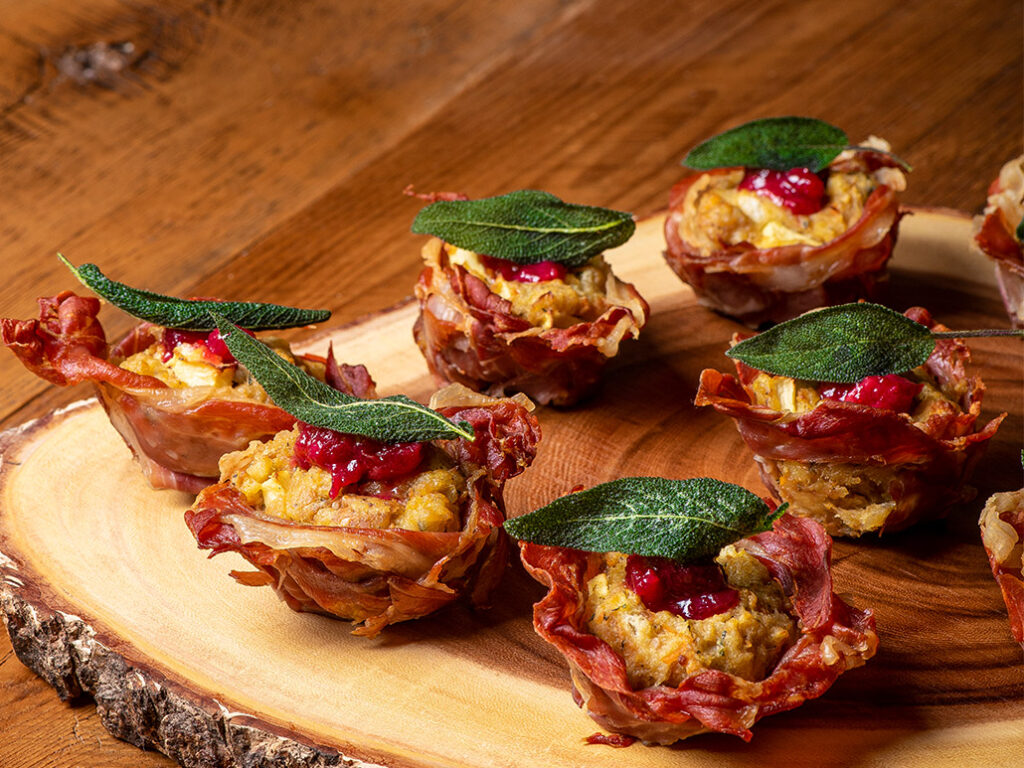 Mini savory cups made of crispy prosciutto, filled with stuffing, topped with cranberry sauce and a fresh sage leaf—perfect for Thanksgiving catering and beautifully arranged on a rustic wooden serving platter.