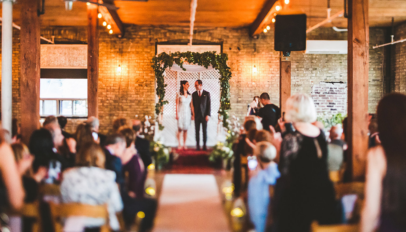 A bride and groom stand together at the altar, smiling, in a warmly lit indoor venue with guests seated on both sides watching the wedding ceremony and savoring exquisite wedding catering.