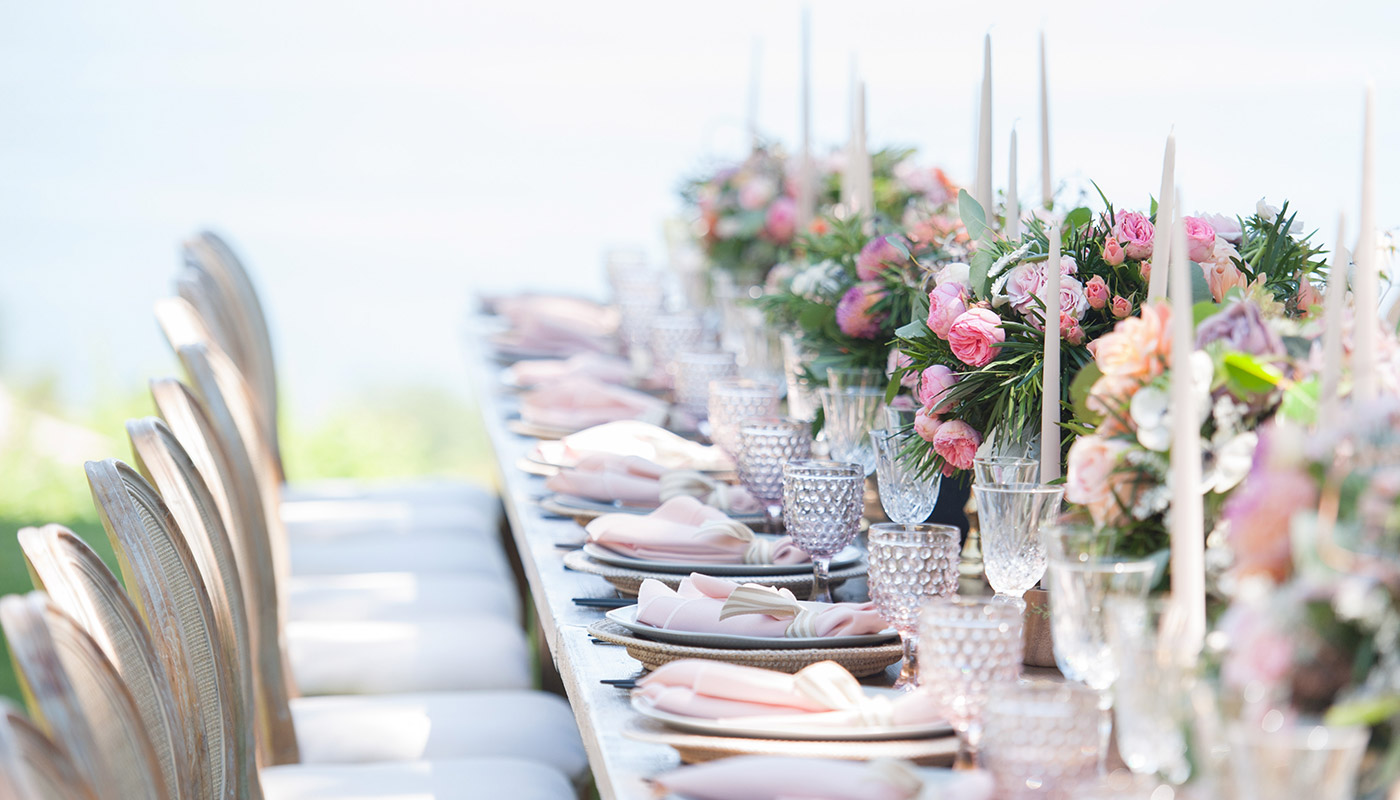 A long, elegant table set outdoors for wedding catering features white chairs, pink napkins, crystal glassware, tall white candles, and lush floral centerpieces with pink and peach flowers.