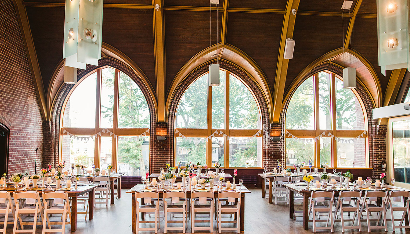 A spacious banquet hall with tall arched windows, wooden beams, and brick walls. Long tables set for a formal event feature elegant wedding catering displays and fresh flowers, with white chairs arranged neatly around them.