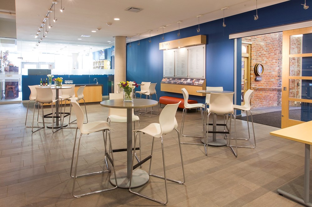 A modern café area with white chairs and round tables on a gray tile floor, blue and brick accent walls, large menu board, and vases of flowers on tables. Natural light comes through glass doors and windows.