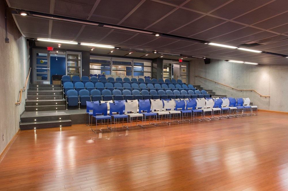 A modern, empty auditorium with wooden floors, grey walls, blue upholstered seats arranged in tiers, and a front row of white and blue chairs on the ground level. Stairs and metal railings are on each side.