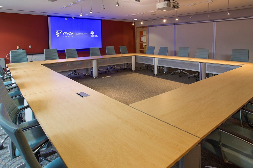A large, modern conference room with a U-shaped arrangement of wooden tables surrounded by blue office chairs. A projection screen displays the YWCA Toronto logo on a red accent wall at the front of the room.