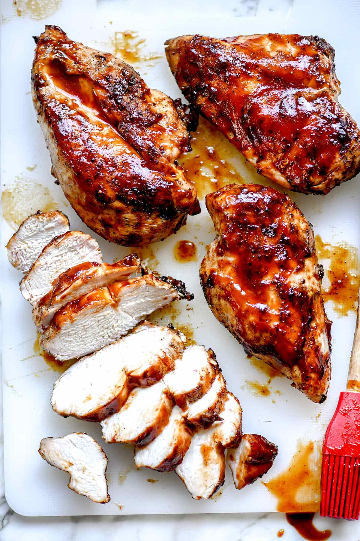 Grilled barbecue chicken breasts, some whole and some sliced, are arranged on a white cutting board with a red basting brush and a drizzle of barbecue sauce.