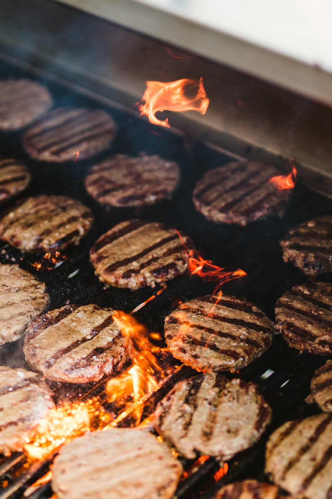 Rows of beef patties with grill marks are cooking on a barbecue grill, with small flames and smoke rising between them.