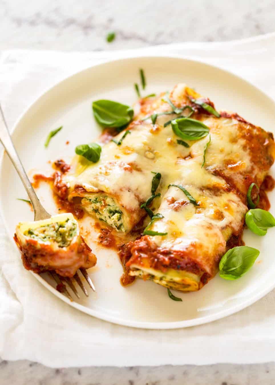A plate of cheesy baked cannelloni topped with tomato sauce and fresh basil leaves, with a fork holding a bite-sized piece showing the green ricotta and spinach filling.