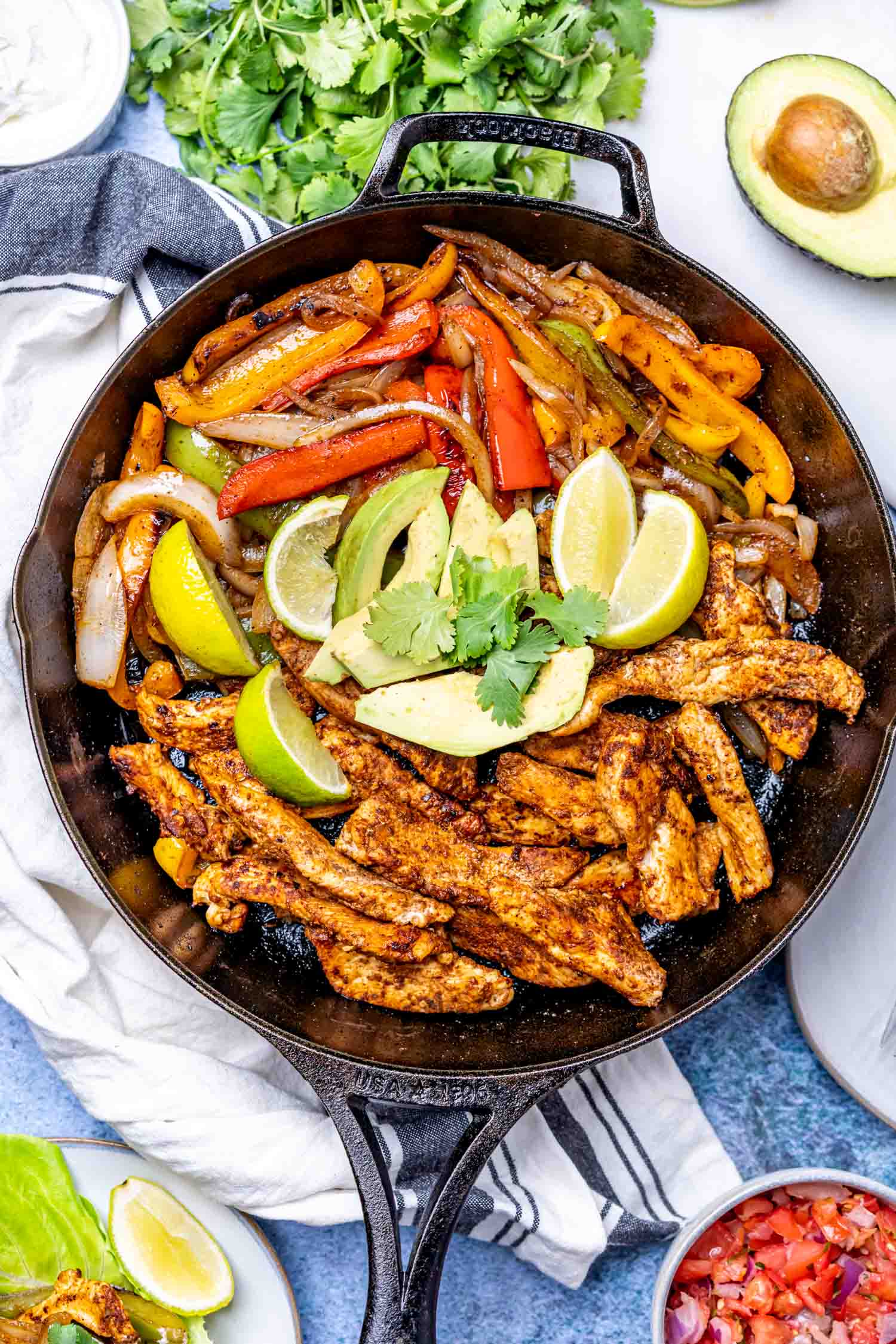 A cast-iron skillet filled with seasoned chicken strips, sautéed red, yellow, and orange bell peppers, onions, avocado slices, lime wedges, and cilantro, surrounded by fresh ingredients on a table.