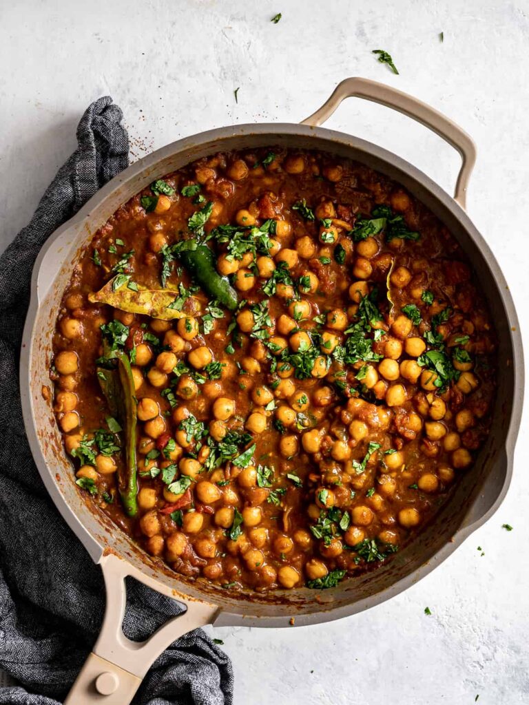 A beige skillet filled with chickpea curry garnished with chopped cilantro sits on a light surface next to a dark kitchen towel. The curry is rich, thick, and features whole spices and green chilies.