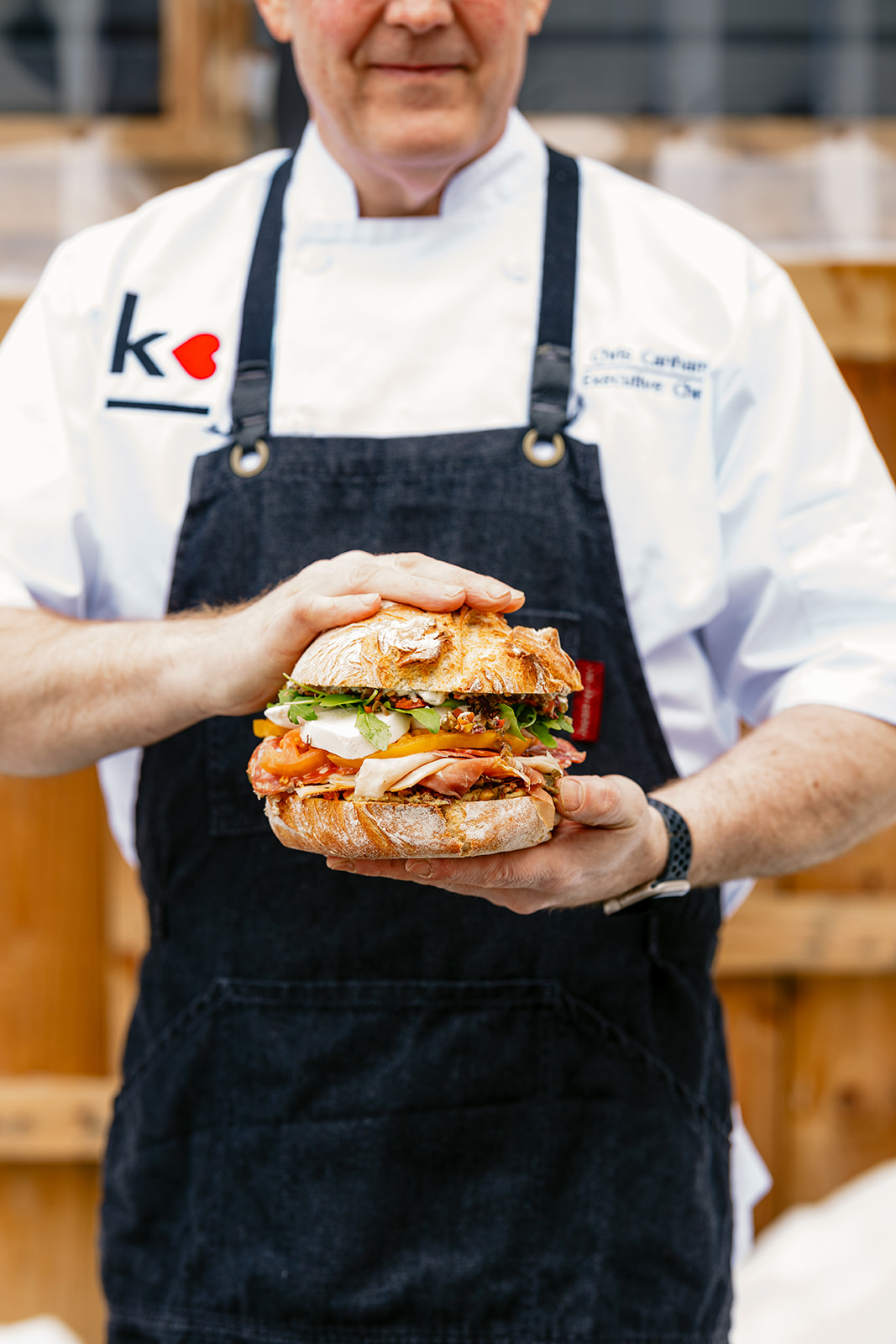 A chef in a white coat and dark apron holds a large sandwich loaded with lettuce, tomatoes, onions, and meat, showing it proudly with both hands. The chef’s face is partially out of frame.