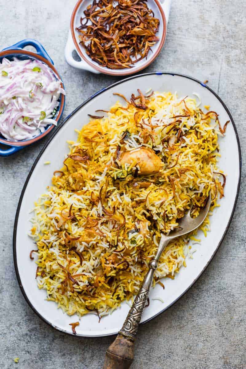 An oval platter of chicken biryani garnished with crispy fried onions, with a serving spoon on the side. Nearby are small bowls of fried onions and sliced onion raita. The dishes are set on a light-colored surface.