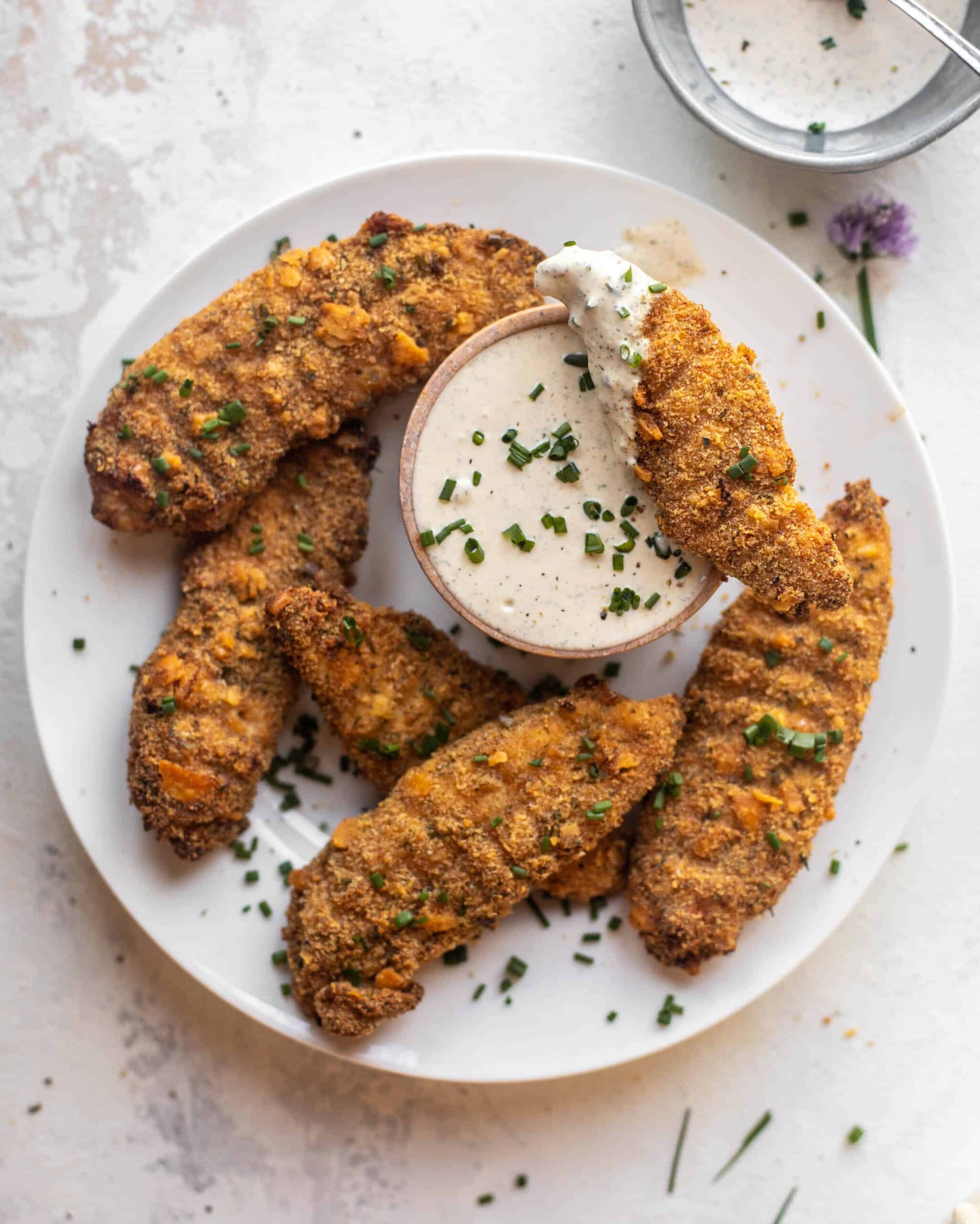 A white plate with five crispy breaded chicken tenders garnished with chopped chives, arranged around a small bowl of creamy dipping sauce. One tender is dipped in the sauce.