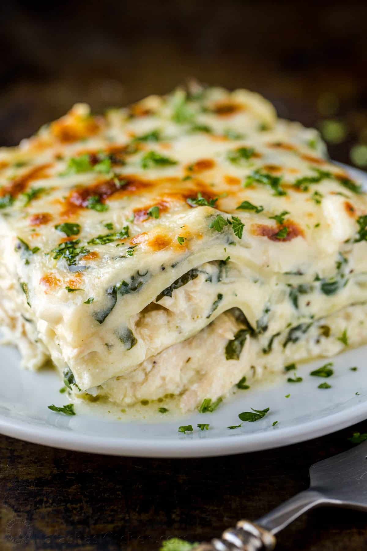 A slice of creamy white lasagna with layers of pasta, spinach, cheese, and béchamel sauce, garnished with chopped parsley, served on a white plate.