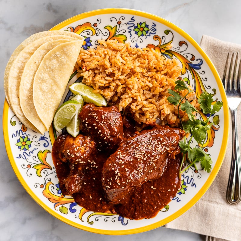 A colorful plate with chicken in dark red mole sauce topped with sesame seeds, served with Mexican rice, corn tortillas, lime wedges, and fresh cilantro. A fork and knife are placed on a beige napkin beside the plate.
