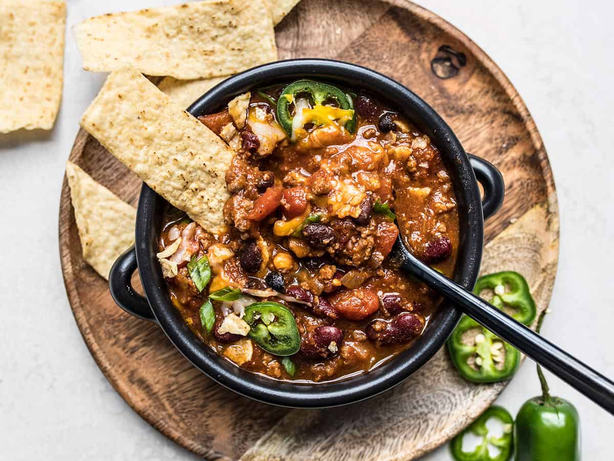 A black bowl of chili topped with sliced jalapeños and green onions, served with tortilla chips on the side. The bowl sits on a round wooden platter, with extra chips and sliced jalapeños nearby.