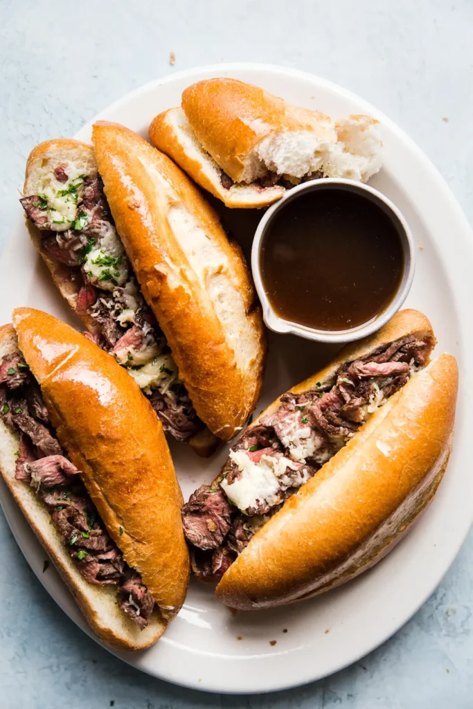 Five French dip sandwiches filled with sliced roast beef and cheese are arranged on a white plate with a small bowl of dark dipping sauce. One sandwich is partially open, revealing its contents.