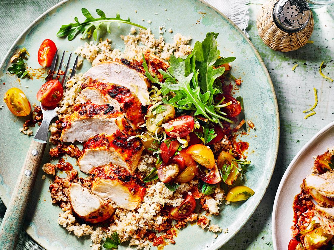 A plate with sliced grilled chicken on a bed of couscous, topped with mixed cherry tomatoes and fresh arugula, with a fork on the side and a pepper shaker in the background.