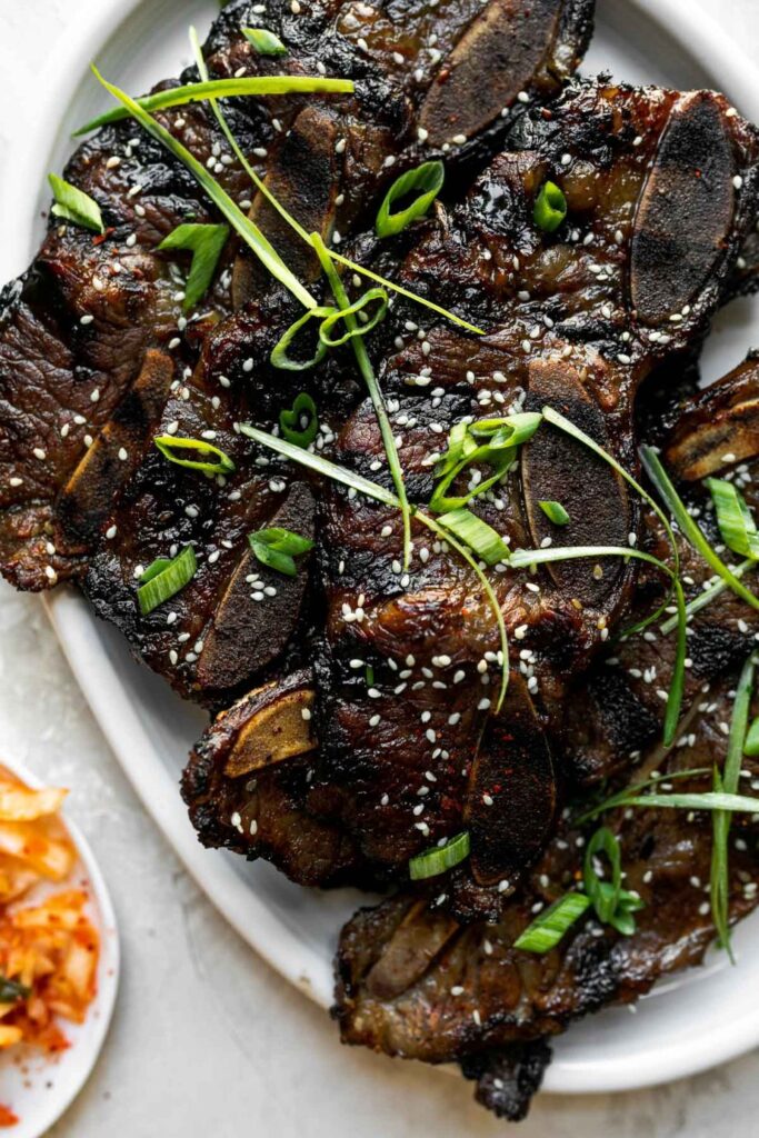 A plate of grilled beef short ribs garnished with sesame seeds and sliced green onions, with a small side of kimchi visible in the corner.