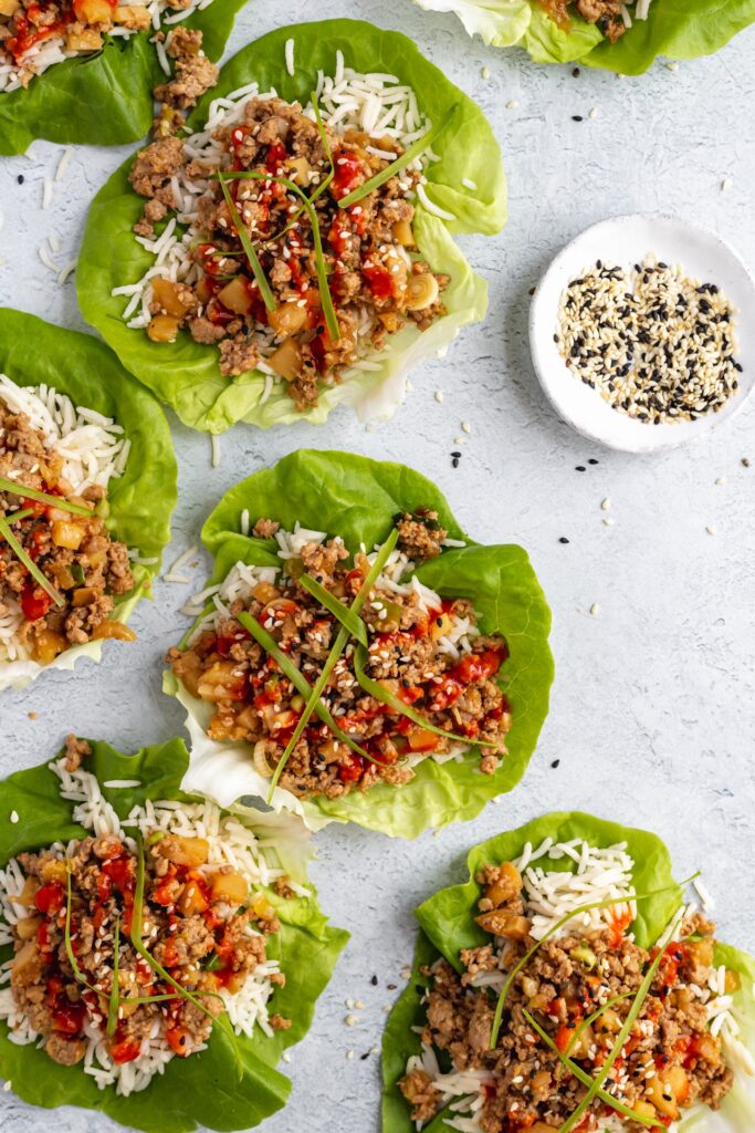 Eight lettuce wraps filled with rice, seasoned ground meat, diced vegetables, and garnished with sesame seeds and chives are arranged on a light surface next to a small bowl of mixed seeds.