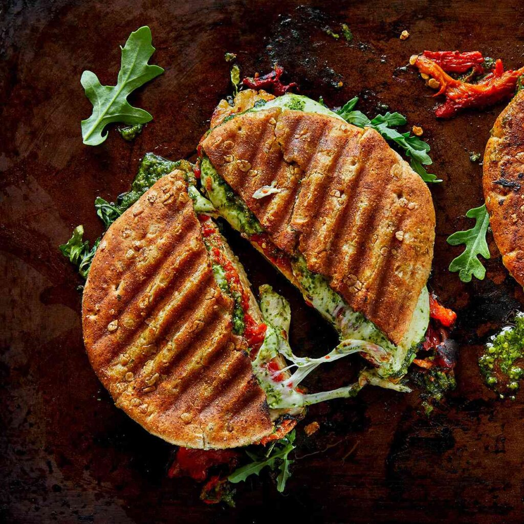 A grilled panini sandwich filled with melted cheese, sun-dried tomatoes, pesto, and fresh arugula, sliced in half with cheese stretching between the halves, on a dark rustic surface.