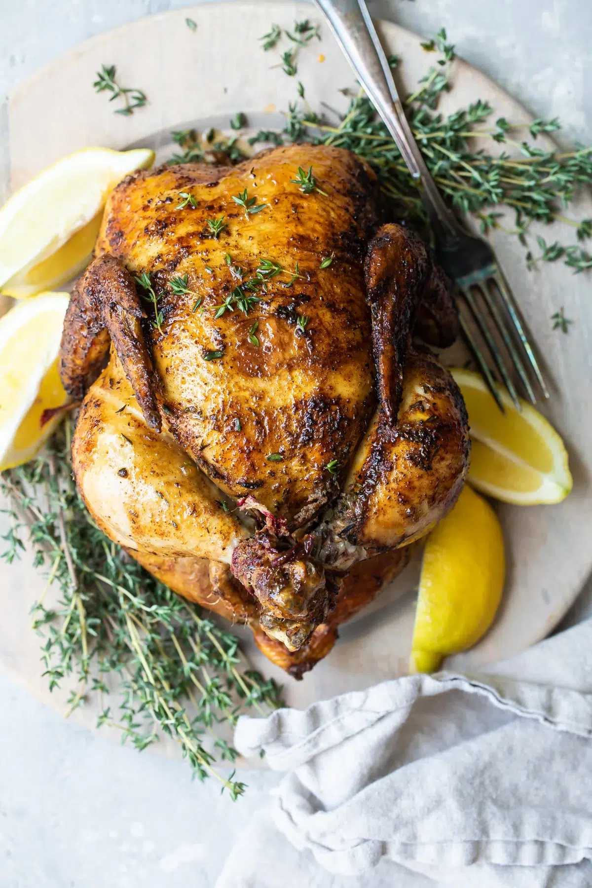 A whole roasted chicken garnished with fresh thyme and lemon wedges sits on a round wooden board, next to a gray napkin and silverware. The chicken has golden, crispy skin.