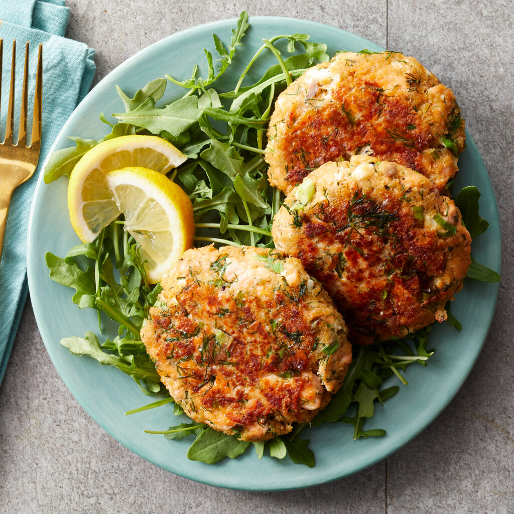 Three golden-brown salmon patties on a bed of arugula, garnished with fresh dill and lemon wedges, served on a light blue plate with a gold fork and teal napkin beside it.
