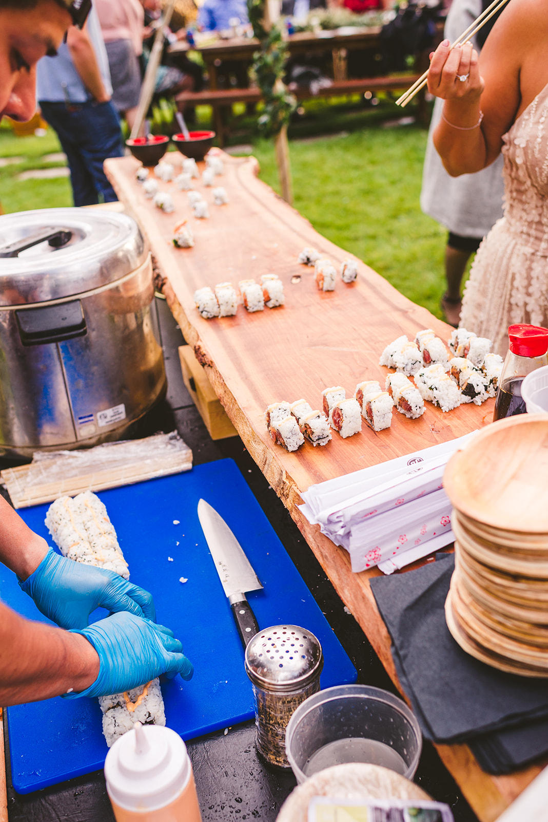 A person wearing blue gloves slices a sushi roll on a blue cutting board at an outdoor food stall, with many pieces of sushi arranged on a wooden serving board and people nearby.