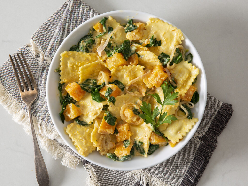 A white bowl filled with ravioli, sautéed spinach, cubes of roasted butternut squash, and onions, garnished with a sprig of parsley. The bowl sits on a folded linen napkin next to a fork.