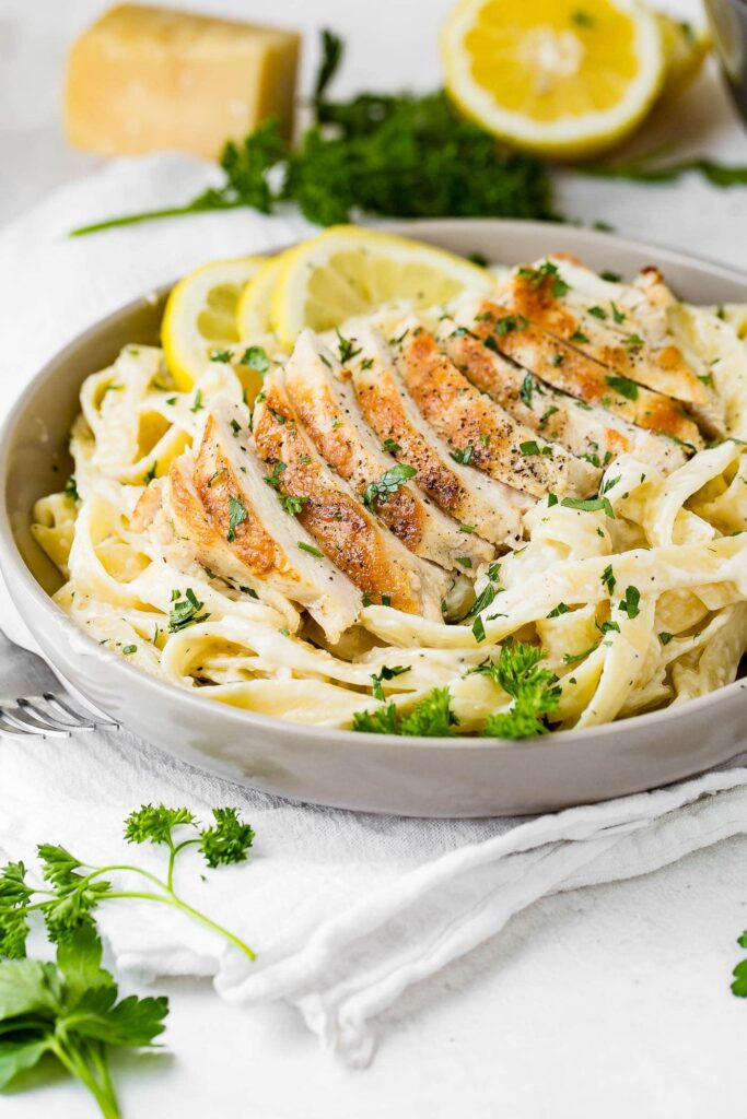 A bowl of creamy fettuccine Alfredo topped with sliced grilled chicken breast, garnished with fresh parsley and lemon slices, with cheese and herbs in the background.