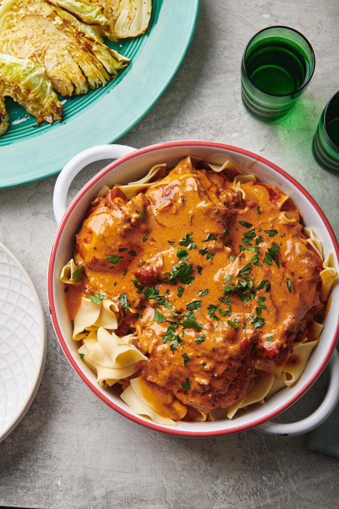 A white dish filled with egg noodles and pieces of chicken covered in a creamy reddish-orange sauce, garnished with chopped parsley. Nearby are green glasses and a plate with roasted cabbage wedges.