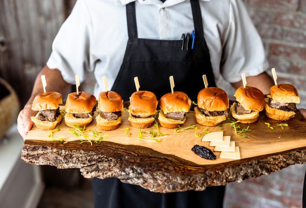 A person in a white shirt and black apron holds a rustic wooden board with seven beef sliders on buns, each with a skewer, garnished with greens and served with cheese slices and a dollop of sauce.
