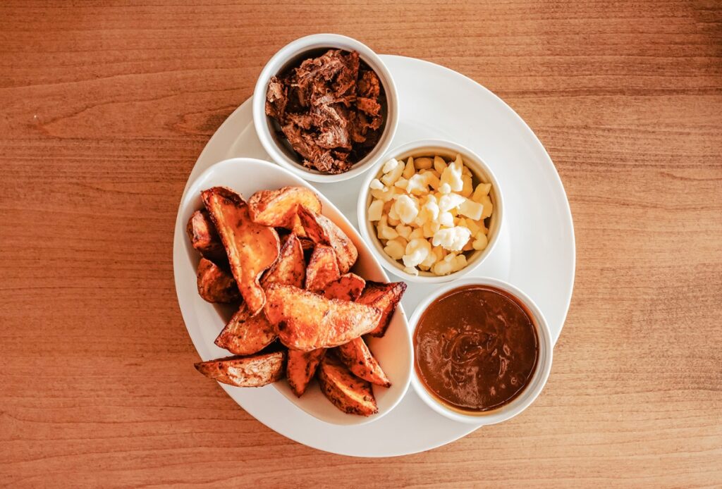 A white plate with potato wedges, shredded meat, cheese curds, and gravy in separate bowls, arranged on a wooden table.