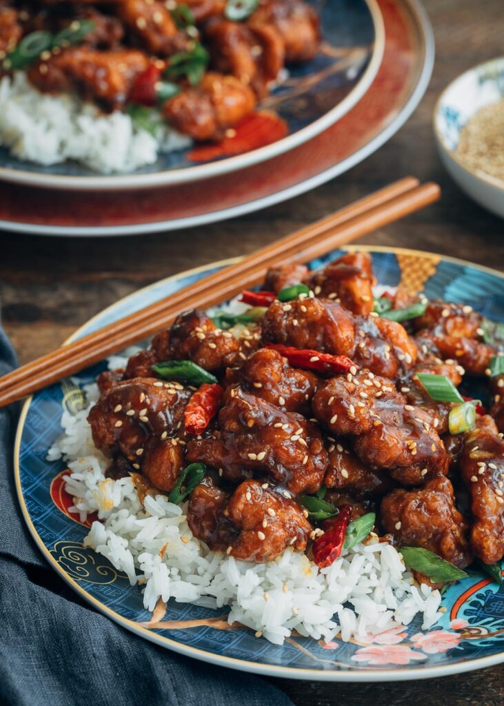 A plate of white rice topped with crispy, saucy pieces of chicken, garnished with sesame seeds, sliced green onions, and red chili peppers, with chopsticks resting on the plate. Another similar plate is in the background.