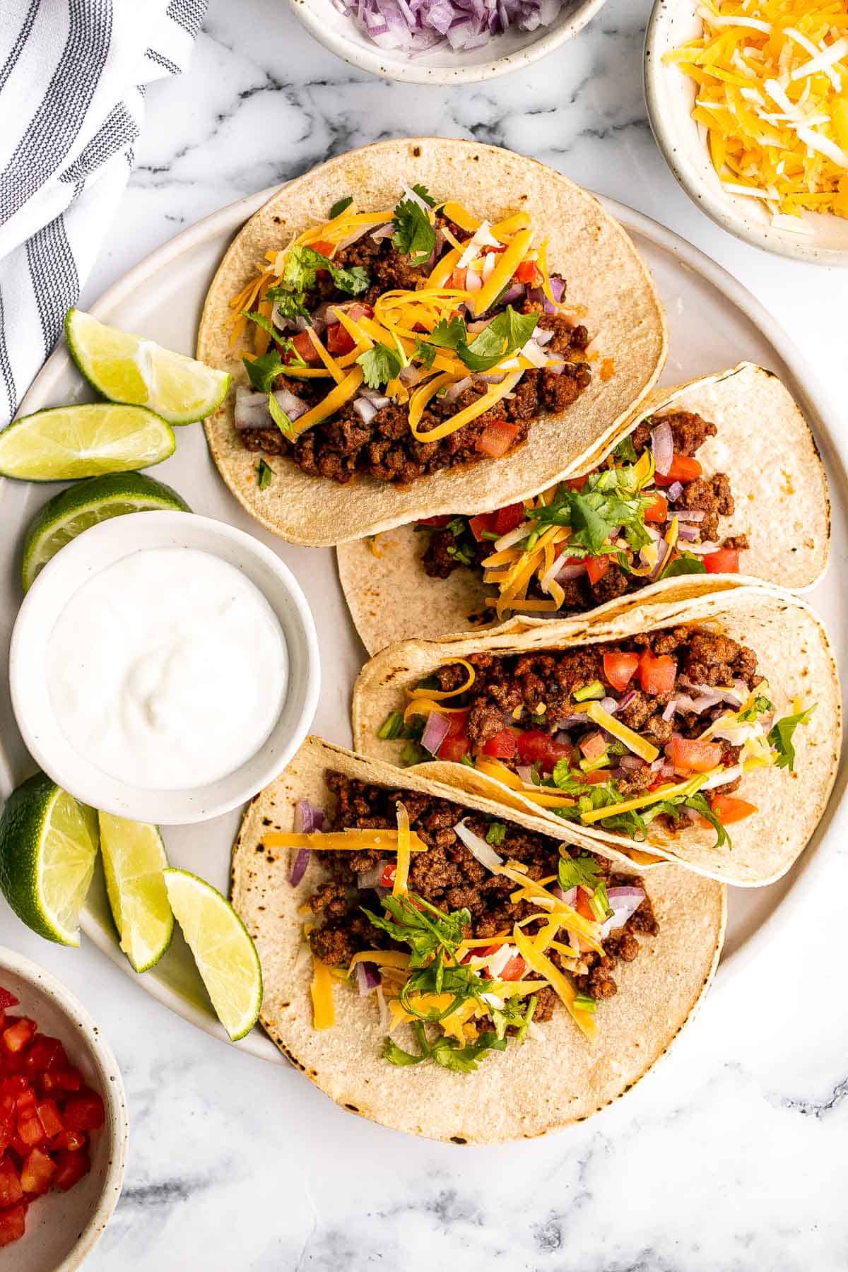 Three soft tacos filled with seasoned ground beef, shredded cheese, diced tomatoes, red onions, and cilantro are on a plate, surrounded by lime wedges, a bowl of sour cream, and extra toppings on a marble surface.