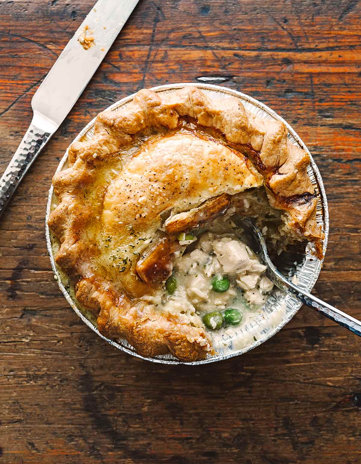 A round chicken pot pie with a golden, flaky crust sits in a foil tin on a wooden table. A portion has been cut out, revealing creamy filling with chicken and green peas. A spoon and knife are beside the pie.