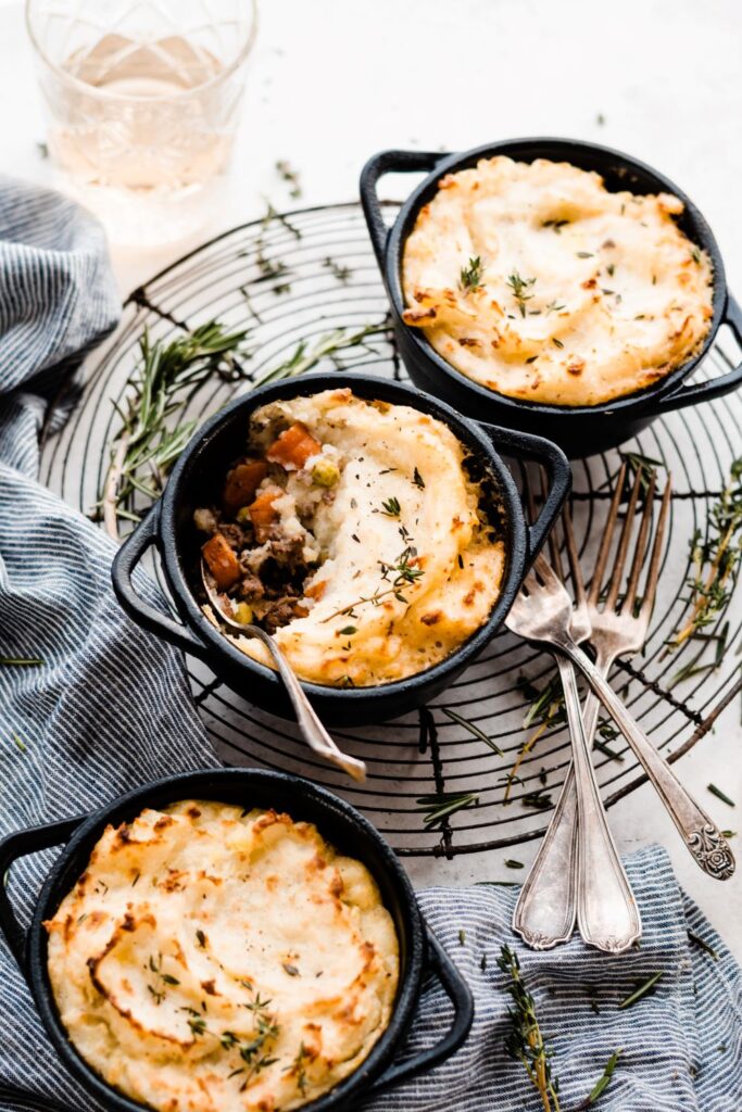 Three mini cast iron pots filled with shepherd’s pie, topped with golden mashed potatoes and garnished with herbs, sit on a wire rack with cutlery and a striped cloth nearby. One pot has a serving scooped out.