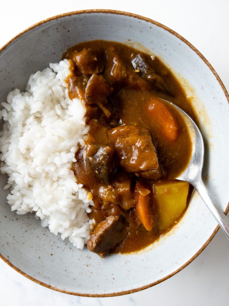 A bowl of white rice served with Japanese curry containing chunks of beef, carrots, and potatoes, with a spoon resting in the curry.