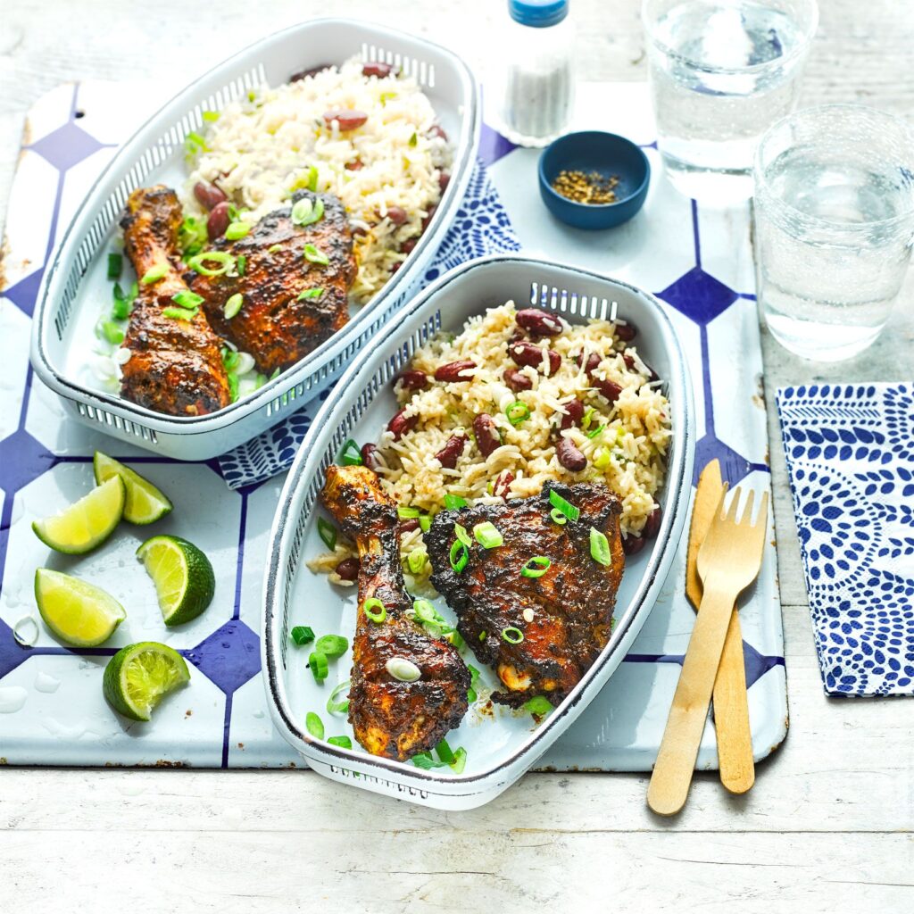 Two trays each contain a grilled chicken drumstick with rice and kidney beans, garnished with chopped green onions. Lime wedges, a fork and knife, water glasses, napkins, and seasonings are set beside the trays.