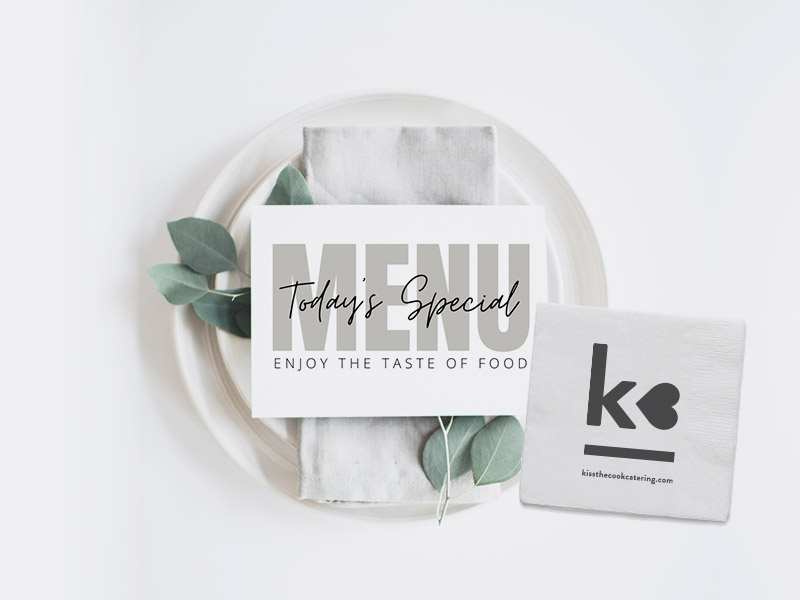 A white plate with a gray napkin and green leaves holds a card reading "MENU Today's Special Enjoy the Taste of Food." Next to the plate is a napkin with "kø" and a website printed on it.