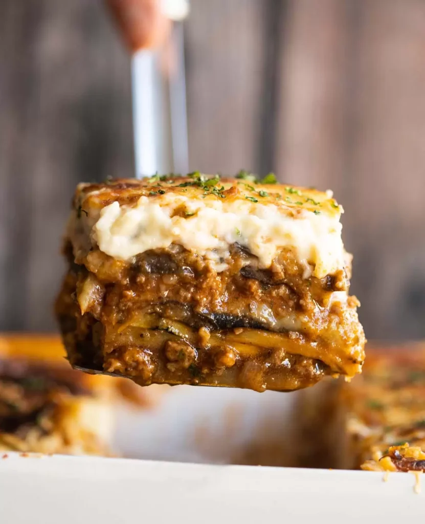 A close-up of a spatula lifting a slice of cheesy, layered lasagna with meat sauce and creamy topping, above a baking dish with more lasagna, against a blurred background.