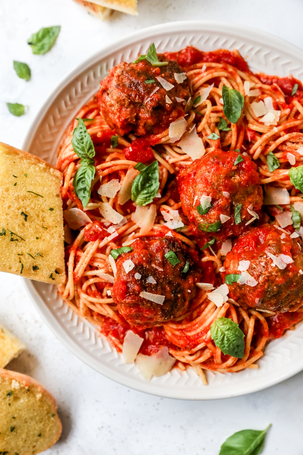 A plate of spaghetti with tomato sauce, four meatballs, fresh basil leaves, shaved Parmesan cheese, and slices of garlic bread on the side.