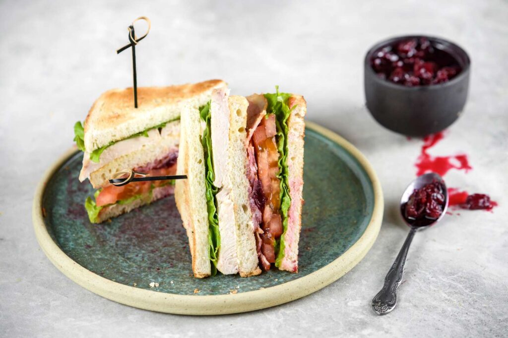 Two turkey club sandwich halves with lettuce, tomato, and cranberry sauce are served on a green plate. A small bowl of cranberry sauce and a spoon with some sauce sit beside the plate on a light surface.