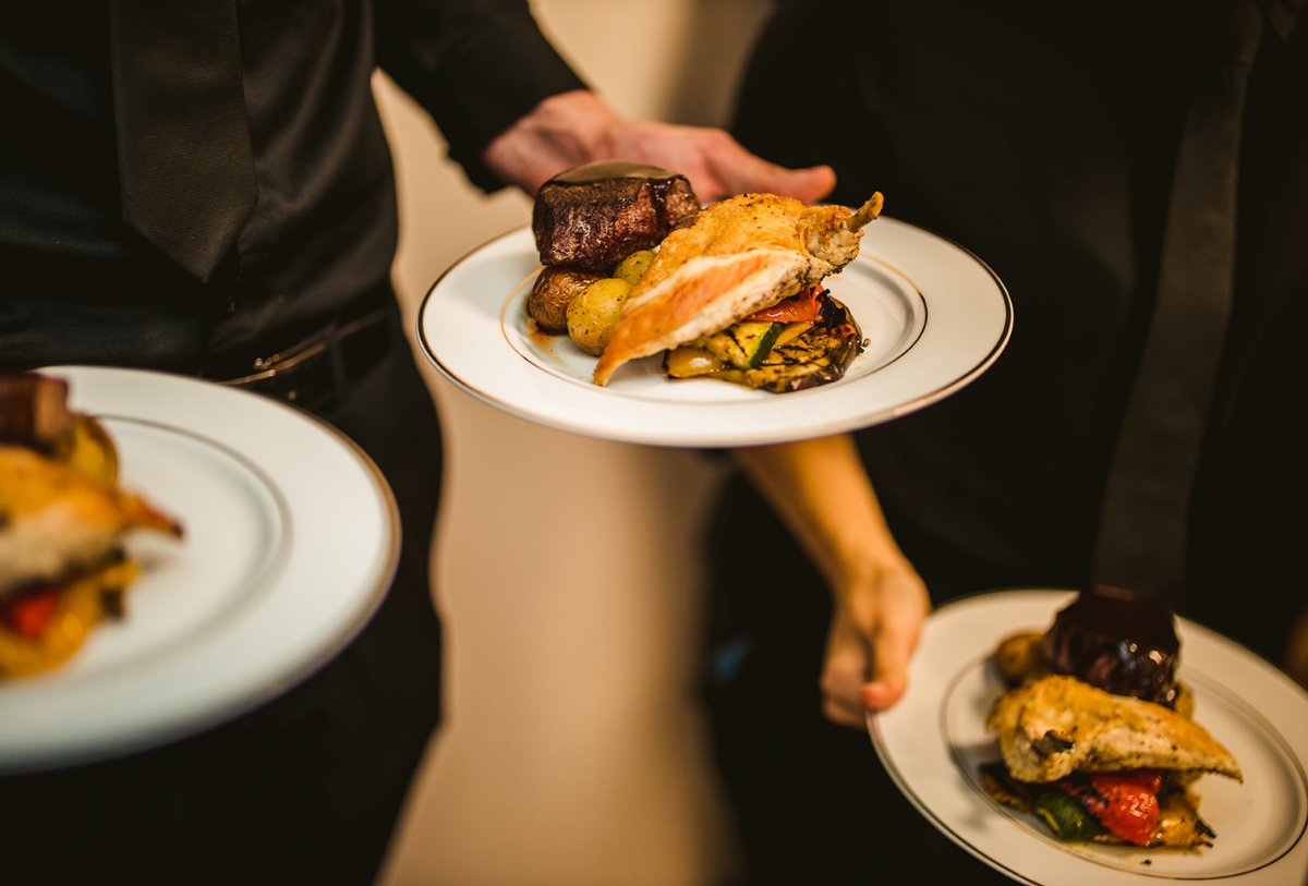 Three servers in black attire hold white plates with gourmet meals, featuring grilled chicken, roasted vegetables, potatoes, and a portion of steak. The focus is on the food, with the servers mostly out of frame.
