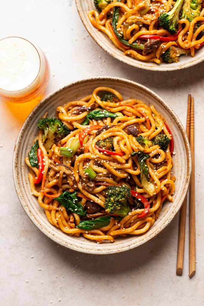A bowl of stir-fried udon noodles with broccoli, red bell pepper, bok choy, and mushrooms, garnished with sesame seeds. Chopsticks rest beside the bowl, and a glass of beer is in the background.